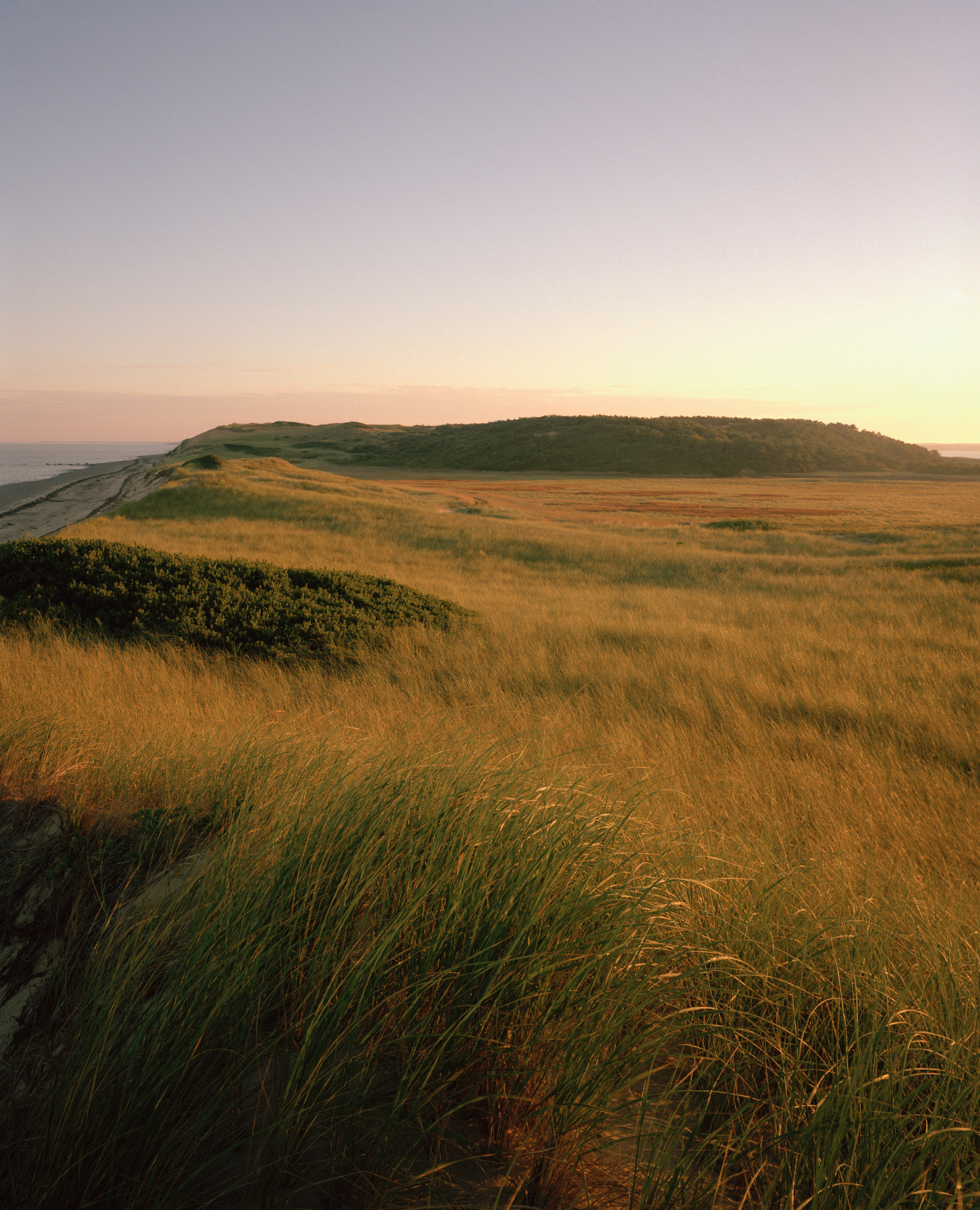 This seven-panel panoramic Cape Cod beach landscape photograph shows the light from a sunrise falling across a spit of beach projecting into a bay.