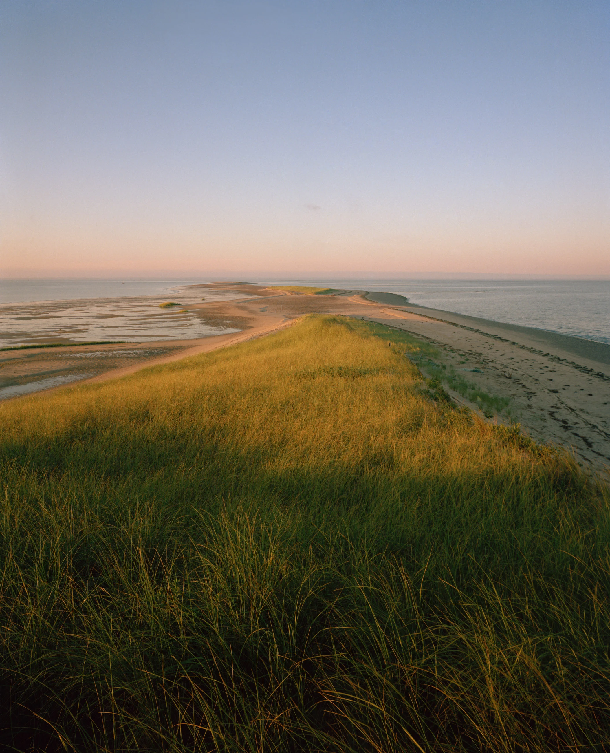 This seven-panel panoramic Cape Cod beach landscape photograph shows the light from a sunrise falling across a spit of beach projecting into a bay.