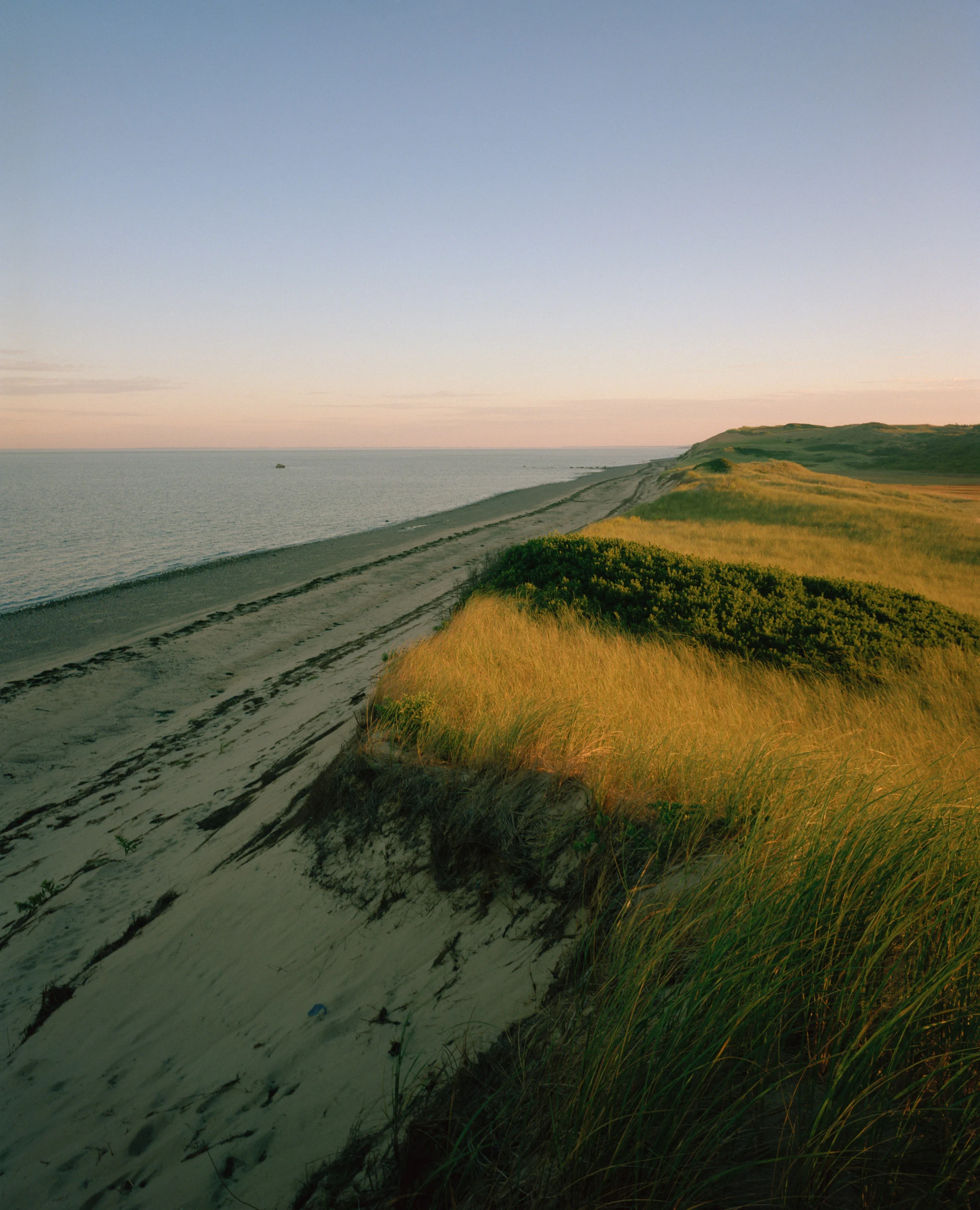This seven-panel panoramic Cape Cod beach landscape photograph shows the light from a sunrise falling across a spit of beach projecting into a bay.